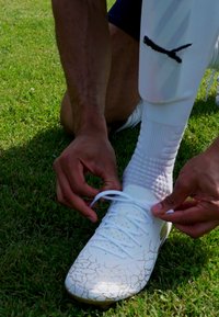 Chaussette de soccer blanche avec un motif craquelé, lacée fermement sur un pied au-dessus d'une pelouse verte. Accompagnée de chaussettes blanches et d'un protège-tibias texturé.