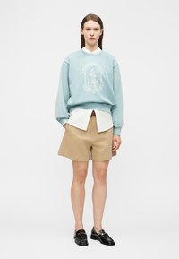 Woman wearing light blue sweatshirt over white shirt, beige knee-length shorts, and black loafers, standing against plain white background.