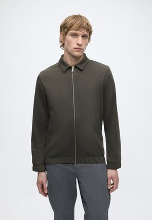 Junger Mann mit kurzen blonden Haaren, trägt eine dunkelgrüne Zip-Jacke und graue Hose, steht vor einem schlichten weißen Hintergrund.