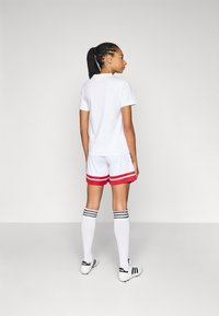 Witte korte mouwen t-shirt, rood en witte sportshorts met strepen, witte kniehoge sokken met zwarte strepen, zwarte voetbalschoenen.