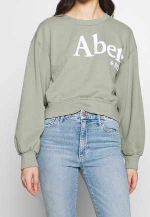 Vrouw draagt een lichtgroene trui met gedeeltelijke witte tekst en lichtblauwe jeans met hoge taille, staand tegen een effen lichte achtergrond.