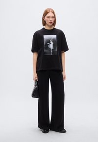 Μαύρο υπερμεγέθες μπλουζάκι με κεντρικό σχέδιο, σε συνδυασμό με μαύρο παντελόνι φαρδιάς γραμμής. Το μοντέλο κρατά ένα μικρό μαύρο τσαντάκι, με λεία υφή.