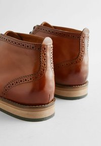 Bottes chelsea en cuir marron avec des détails en brogue, bouts ronds et talons en bois empilés avec une base contrastante.