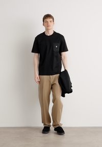 Czarna koszulka z kieszenią na piersi i małym logo, zestawiona z beżowymi spodniami i czarnymi butami, trzymała czarną kurtkę w jednej ręce.