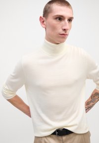 Pull à col roulé crème en tissu doux, avec des manches longues et un design ajusté, porté avec un pantalon beige et une ceinture noire.