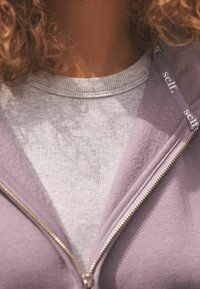 Fialová mikina na zips s rebrovaným šedým tričkom pod ňou. Mikina má strieborný zips a branding na lemovaní. Viditeľné jemné textúry látky.