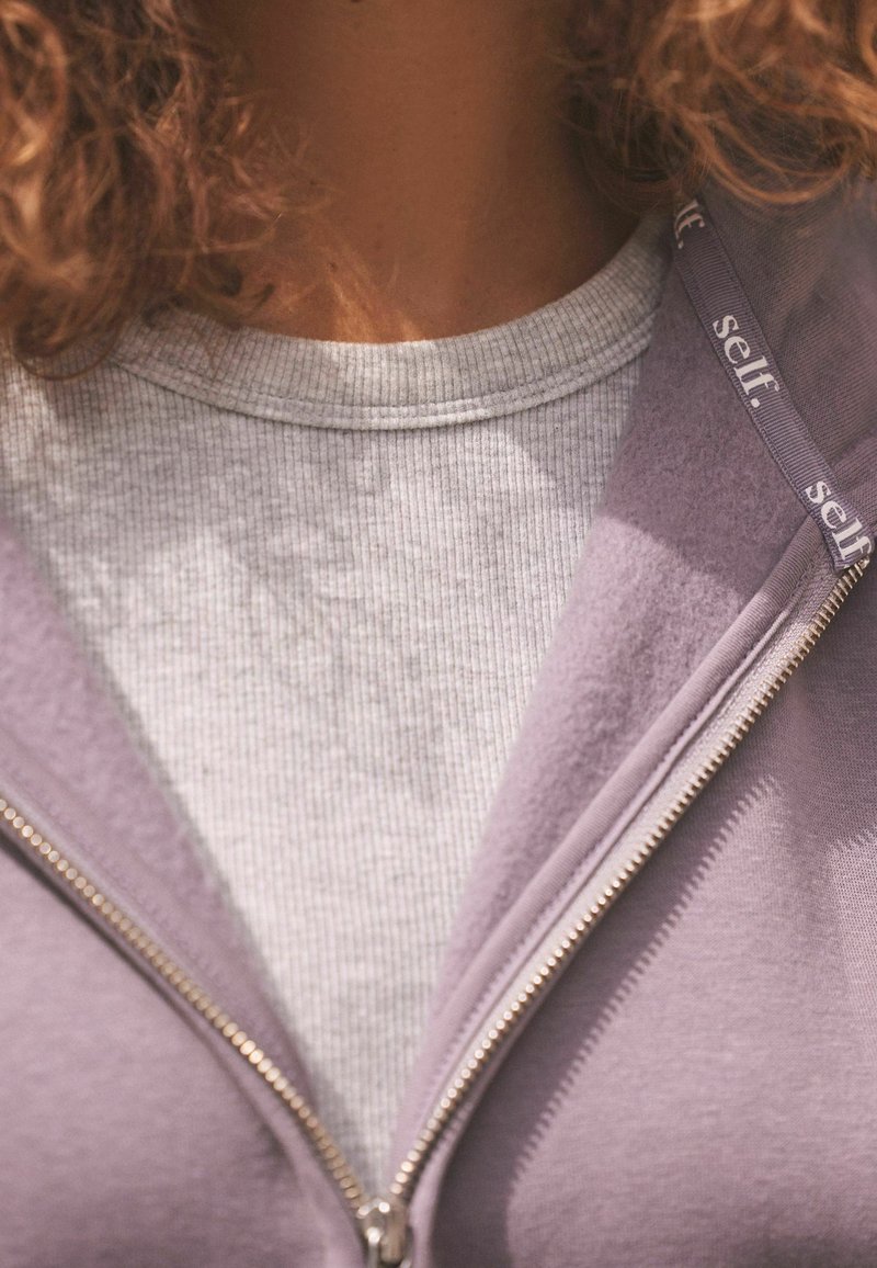 Fialová mikina na zips s rebrovaným šedým tričkom pod ňou. Mikina má strieborný zips a branding na lemovaní. Viditeľné jemné textúry látky.