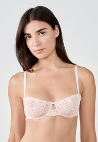 Soutien-gorge en dentelle rose tendre avec des bretelles réglables, présentant un détail en trou de serrure au centre et des bords festonnés sur le haut.