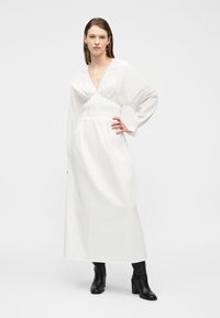 Γυναίκα που φοράει μακρύ λευκό φόρεμα με σούρες στη μέση και V-λαιμόκοψη, μαύρα μπότες με τακούνι, στέκεται με το ένα χέρι στη μέση μπροστά από απλό φόντο.