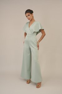 Macacão verde menta com decote em V e mangas flutuantes, apresentando pernas largas e uma cintura ajustada. Usado com sandálias douradas e acessórios.