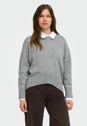 Femme portant un pull gris en maille par-dessus une chemise blanche à col et un pantalon marron foncé, debout devant un fond clair uni.