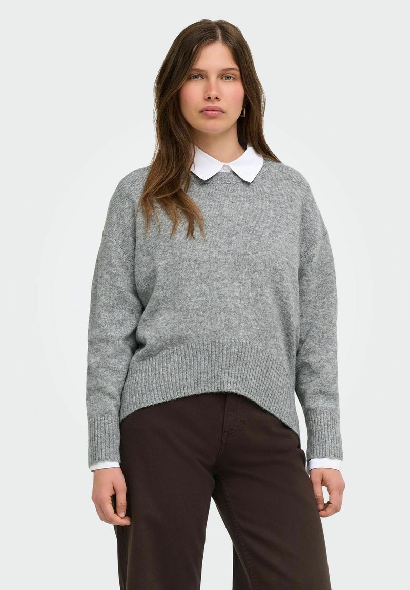 Femme portant un pull gris en maille par-dessus une chemise blanche à col et un pantalon marron foncé, debout devant un fond clair uni.