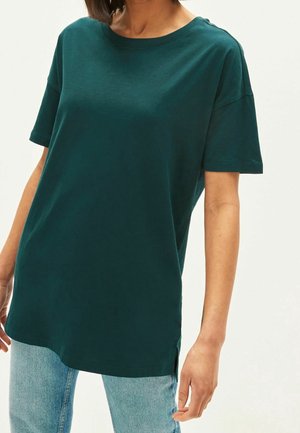 Personne portant un t-shirt ample vert foncé à manches courtes et un jean bleu clair sur un fond clair uni.