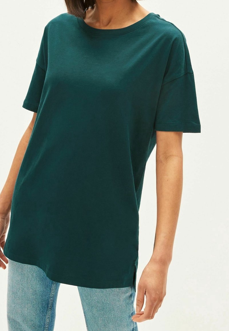Personne portant un t-shirt ample vert foncé à manches courtes et un jean bleu clair sur un fond clair uni.