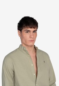 Camisa de botón verde claro con cuello mandarín y pequeño detalle bordado. Presenta una textura suave y un corte relajado.