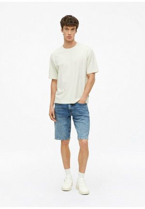 Jonge man die een effen lichtbeige t-shirt draagt, blauwe denim shorts, witte sokken en witte sneakers, staand met één hand in zijn zak.