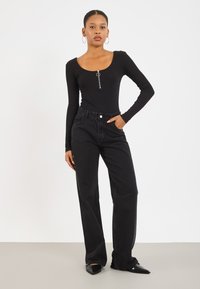Zwarte longsleeve bodysuit met een ronde hals en ritsdetail, gecombineerd met hoge zwarte wijde jeans en puntige zwarte schoenen.