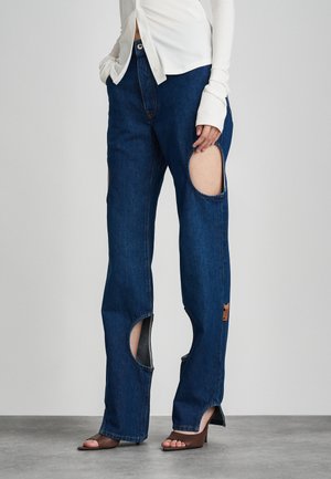 Μπλε τζιν με μεγάλες τρύπες στους μηρούς, ίσια γραμμή, χαμηλή μέση και καφέ δερμάτινη διακόσμηση κοντά στο τελείωμα.