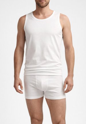 Uomo che indossa una canottiera bianca semplice e boxer bianchi, in piedi contro uno sfondo neutro.