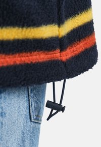Marineblaue Fleecejacke mit Saum in Gelb und Orange, verstellbarer schwarzer Kordelzug mit Stopper, getragen über hellblauen Jeans.