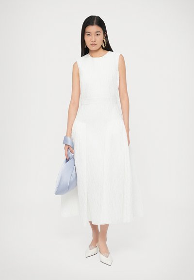 Bela obleka brez rokavov z teksturiranim vzorcem, srednje dolžine, kombinirana z svetlo modro torbico in belimi koničastimi čevlji.