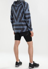 Veste légère à capuche en bleu foncé avec des rayures géométriques noires. Associée à un short noir et des chaussures de sport. Matière lisse et imperméable.