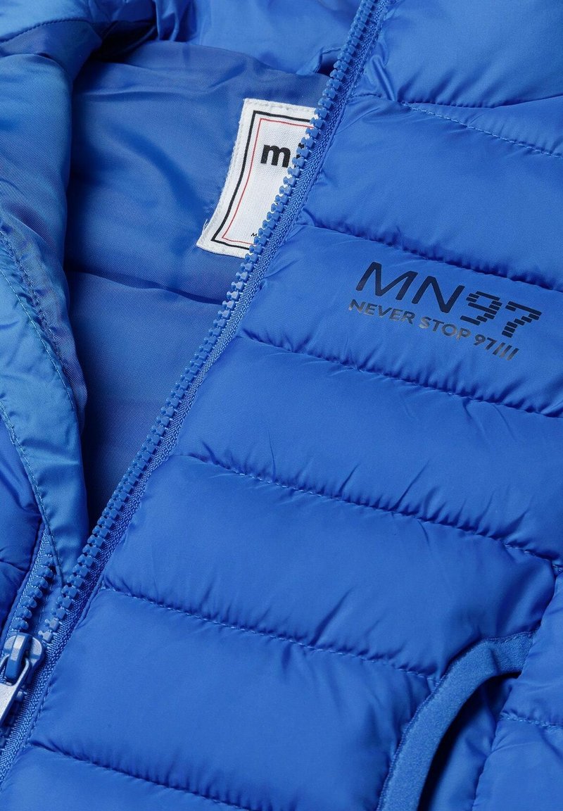 Doudoune bleue avec texture matelassée, dotée d'une fermeture éclair à l'avant et d'une étiquette de marque. Les accents comprennent un logo en relief noir.