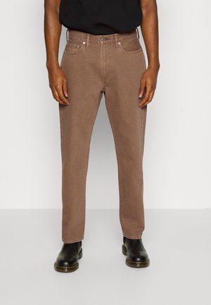 Mann trägt braune Jeans mit geradem Bein, schwarze Lederstiefel und ein schwarzes Hemd, steht vor einem hellgrauen Hintergrund.