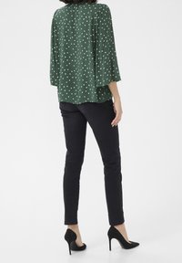 Blusa verde a pois con maniche morbide, abbinata a jeans skinny neri e tacchi alti neri. La blusa ha una vestibilità rilassata e una texture morbida.