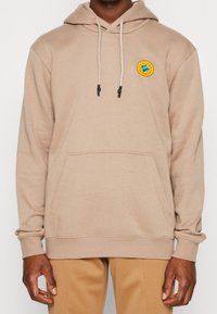 Man som bär en beige huppare med framficka och vita dragsnören, med en liten rund färgglad patch på vänster bröst.