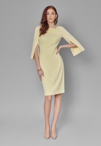 Żółta sukienka o smukłej sylwetce, z rękawami typu dzwon do łokcia, bocznymi rozcięciami i gładkiego materiału. Połączona z cielistymi szpilkami.