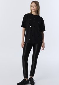 Czarna koszulka z krótkim rękawem, pokryta odblaskowymi kropeczkami, w zestawieniu z błyszczącymi czarnymi legginsami i teksturowanymi czarnymi sneakersami. Minimalistyczny design.