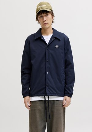 Νεαρός άνδρας που φοράει μπεζ καπέλο, μπλε σκούρο μπουφάν με κουμπιά πάνω από λευκό πουκάμισο και χαλαρό καφέ παντελόνι, κοιτώντας μπροστά με φόντο απλό.