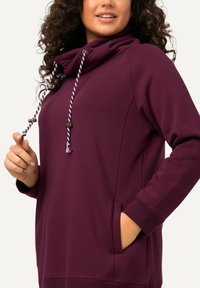 Felpa bordeaux con collo a cowl, cordini regolabili e tasche laterali. Texture morbida con polsini a costine e cordino a contrasto.