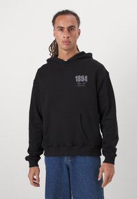 Hoodie noir avec une poche avant, des poignets côtelés et un graphique avec le numéro "1894" en texte gris sur le côté gauche de la poitrine. Porté avec un jean bleu.