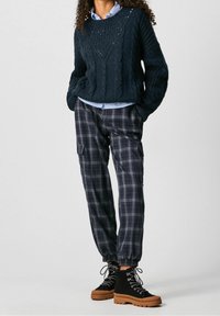 Kobieta w czarnym swetrze z dzianiny w splot warkoczowy, nosząca niebieską koszulę w paski, spodenki cargo w kratę oraz czarne buty na sznurowadła z grubą brązową podeszwą.