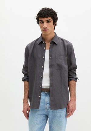 Junger Mann trägt ein aufgeknöpftes dunkelgraues Langarmhemd über einem weißen Tanktop, hellblaue Jeans und einen braunen Gürtel, steht vor einem einfarbigen weißen Hintergrund.