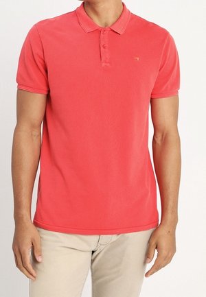 Homme portant un polo rouge corail à manches courtes et un pantalon beige, debout devant un fond blanc uni.