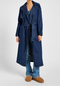 Abrigo de mezclilla en azul oscuro con diseño de doble botonadura, gran cuello y cintura ceñida con cinturón. Lleva puesto sobre unos jeans de color azul claro y calzado color marrón claro.