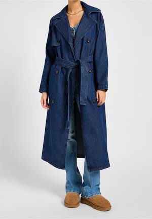 Denim trenčkot v tmavě modré barvě s dvouřadým zapínáním, velkým límcem a páskem v pase. Nosený přes světle modré džíny a béžovou obuv.