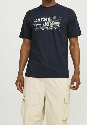 Uomo che indossa una t-shirt grafica "Jack & Jones" blu navy e pantaloncini cargo beige su uno sfondo chiaro e semplice.