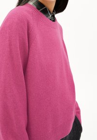 Pull tricoté rose avec un col rond, des poignets et un ourlet côtelés. La texture semble douce ; porté sur une chemise à carreaux.