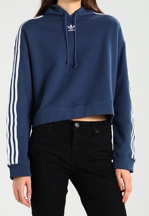 Kobieta ubrana w krótki granatowy kaptur Adidas z białymi paskami na rękawach oraz czarne spodnie z wysokim stanem, stojąca z rękami swobodnie opuszczonymi.