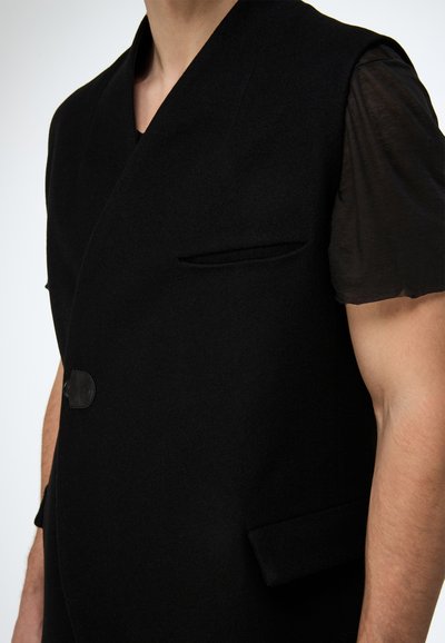 Schwarze Wollweste mit tiefem V-Ausschnitt, Seitentaschen und einem einzelnen Knopfverschluss. Der Stoff hat eine glatte, strukturierte Oberfläche.