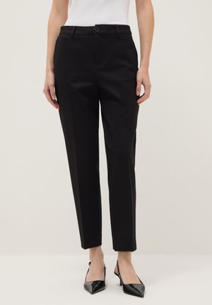 Frau trägt schwarze, maßgeschneiderte knöchellange Hose und schwarze Slingback-Pumps mit spitzer Spitze, steht vor schlichtem Hintergrund.