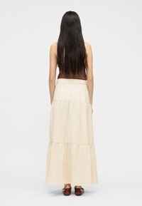 Γυναίκα με μακριά σκούρα μαλλιά, φοράει καφέ αμάνικο τοπ, μαξιμαλιστική φούστα σε κρεμ αποχρώσεις με βολάν και καφέ κορδονιαστά παπούτσια, στέκεται γυρισμένη με την πλάτη.