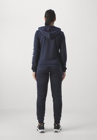 Námořnická modrá mikina a souprava tepláků vyrobená z měkkého materiálu, s kapucí na šňůrku a přiléhavými manžetami, doplněná černými teniskami.