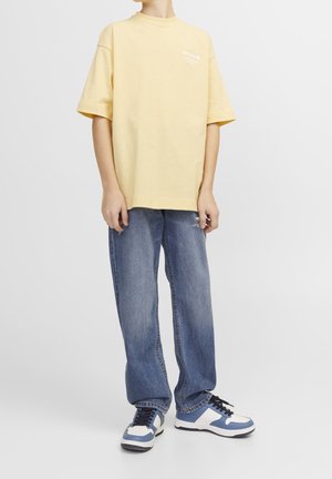 T-shirt di cotone giallo chiaro con maniche corte e colletto rotondo, abbinata a jeans blu a taglio rilassato. Sneakers blu e bianche completano il look.
