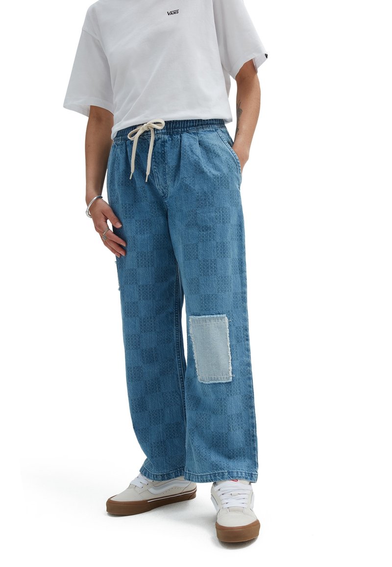 Personne portant un t-shirt Vans blanc, un pantalon large en jean à carreaux bleus avec un patch clair au genou, des baskets blanches avec des semelles marron.