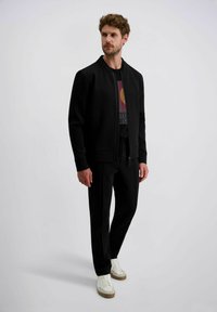 Schwarze Bomberjacke mit Reißverschluss und gerippten Bündchen, kombiniert mit schwarzen Hosen und weißen Sneakers. Das T-Shirt hat ein farbenfrohes Grafikdesign.
